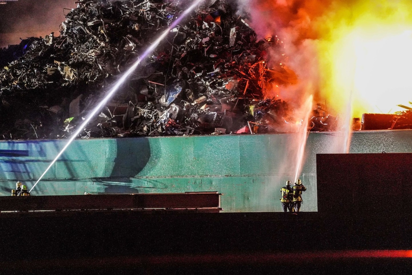 Ein großer Schrotthaufen brennt und wird von Feuerwehrleuten gelöscht.