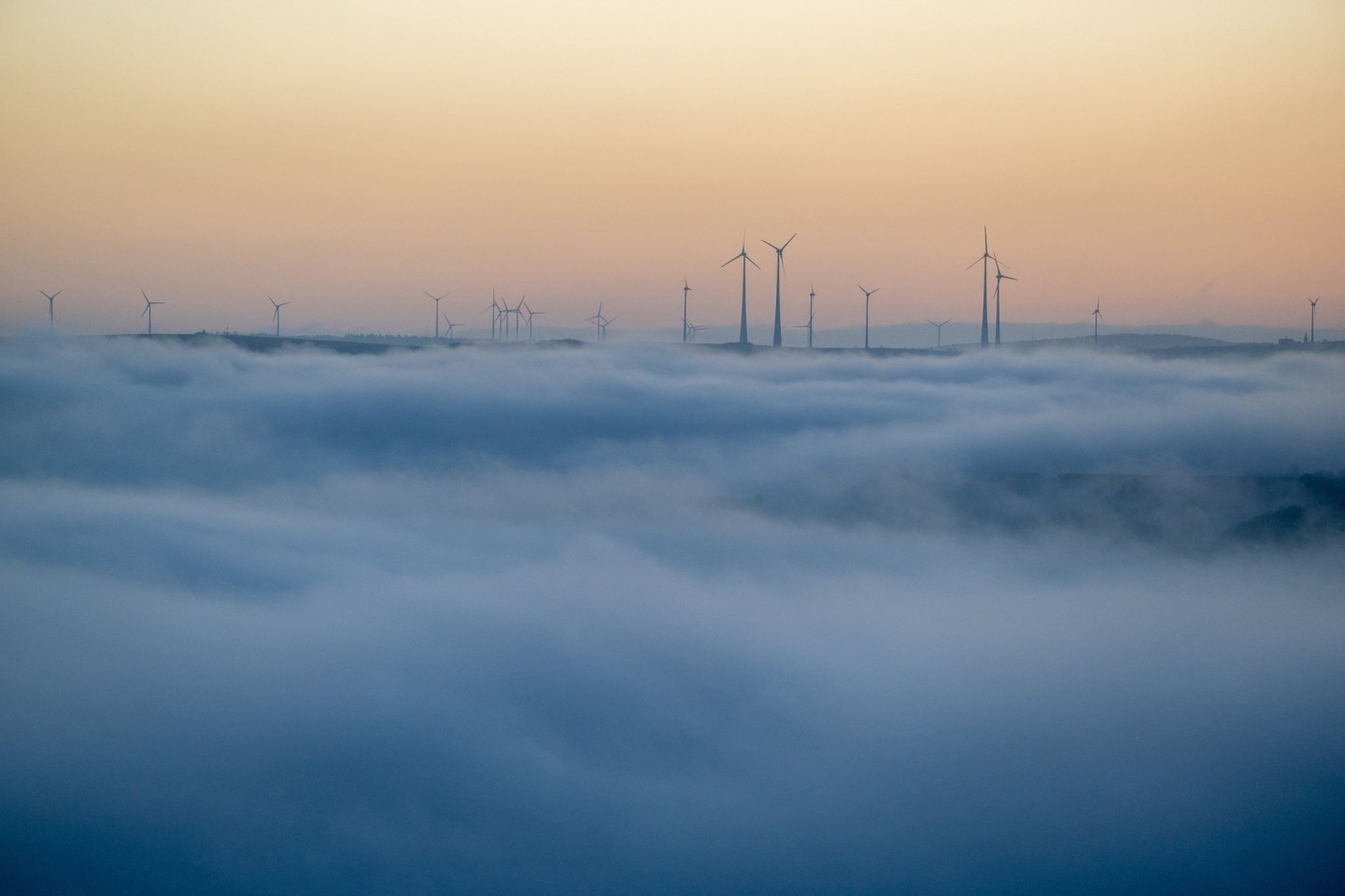 Windkraftanlegen im Hochwald ragen am Morgen aus dem Nebel, unter dem die Stadt Trier noch im Verborgenen liegt.
