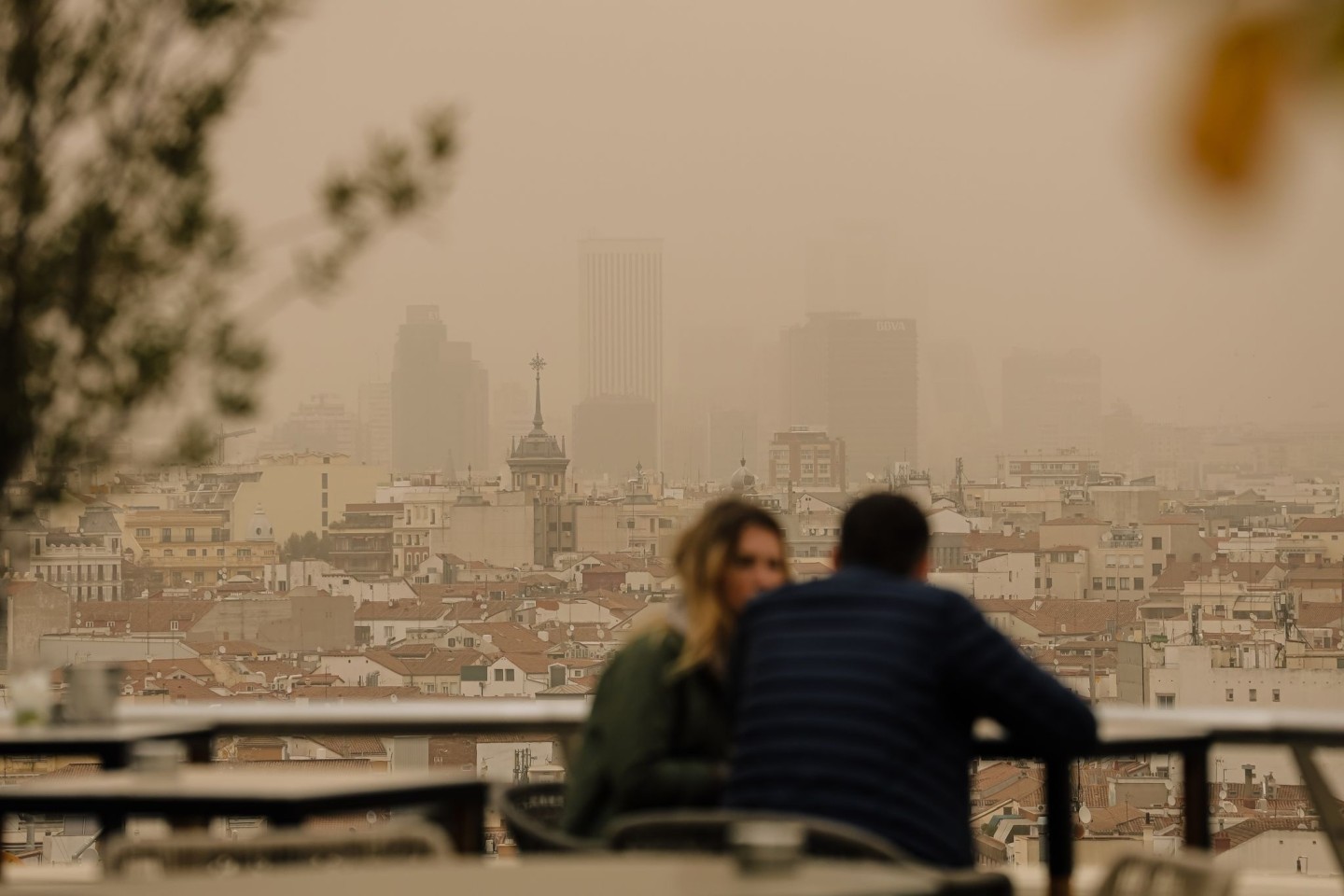 Zwei Menschen sitzen in Madrid auf einer Terrasse, während eine Wolke mit Sahara-Staub über die spanische Hauptstadt zieht. Die typische Auswirkung von Sahara-Staub ist ein roter oder oran...