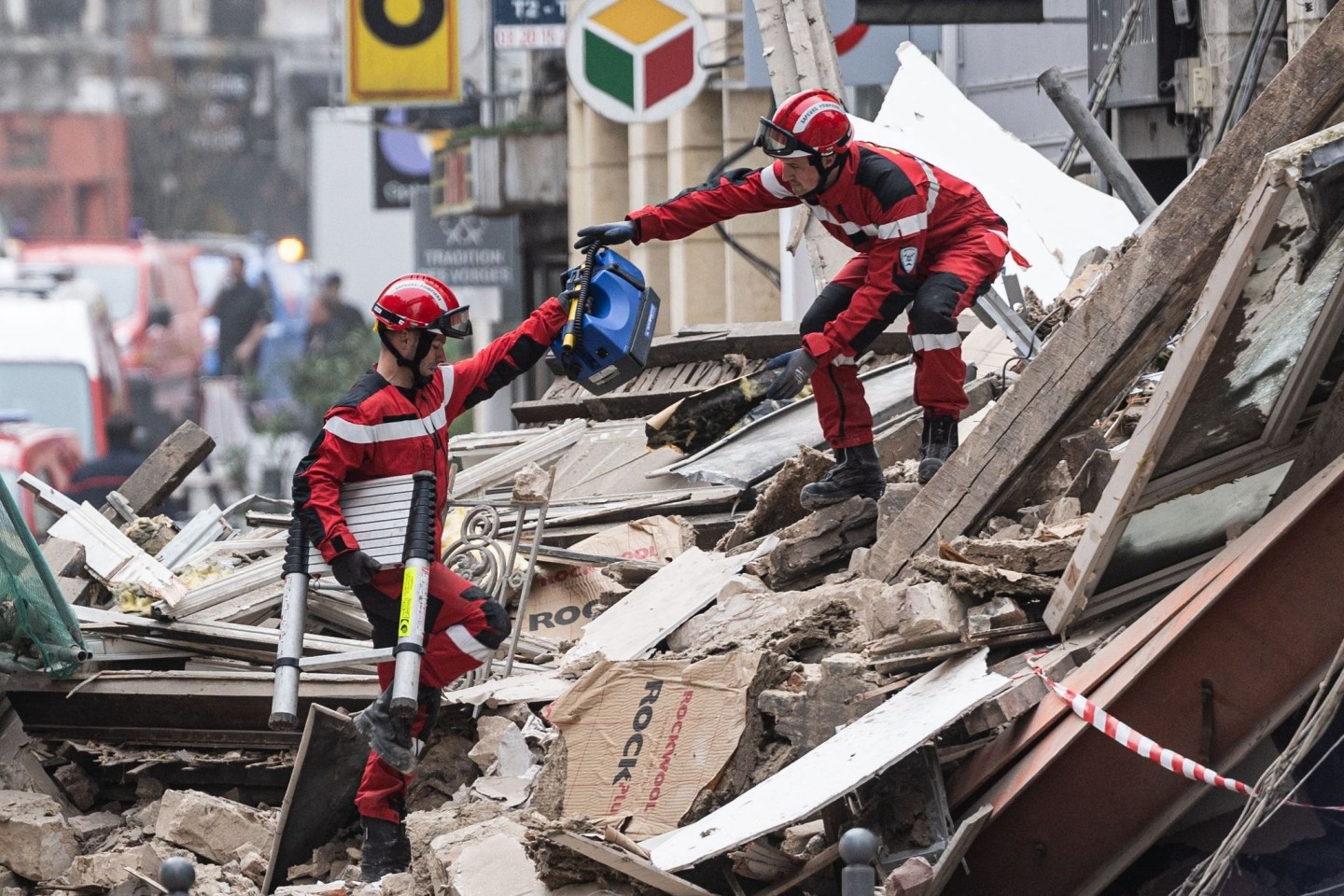 Zwei Rettungskräfte reichen sich Werkzeuge inmitten der Trümmer eines eingestürzten Gebäudes.